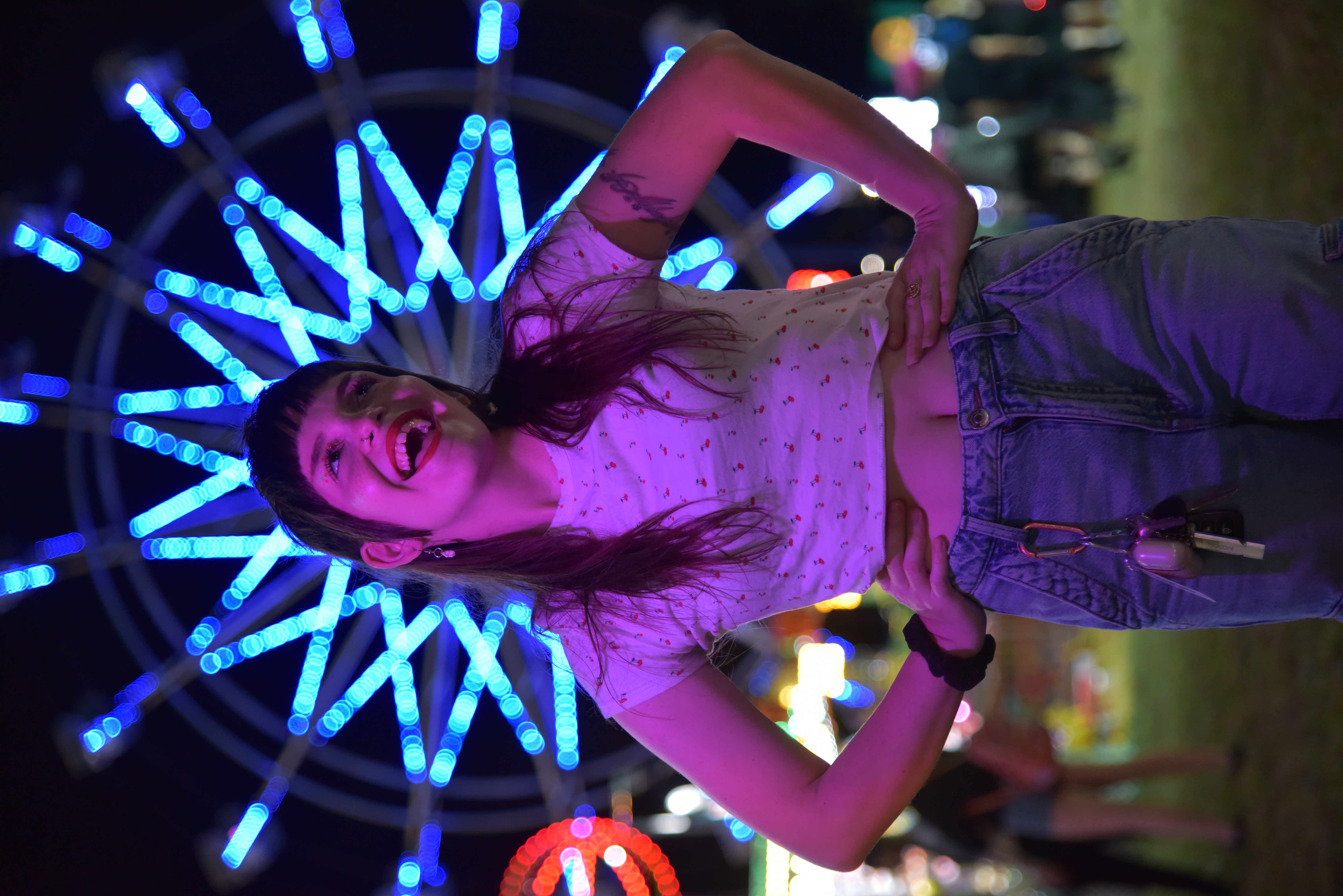 Artist, Alyssa Gilleo smiling in bright red lipstick, jeans, and a cherry shirt with her hands on her hips in front of a Farris Wheel at night, making it look like a crown.
