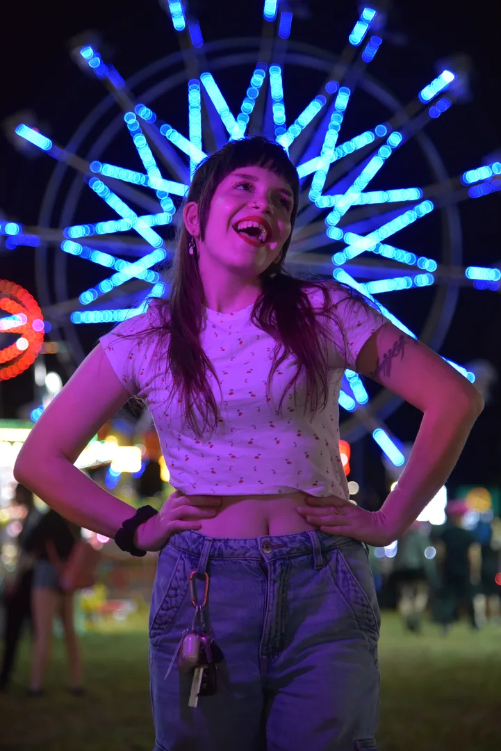 Artist, Alyssa Gilleo smiling in bright red lipstick, jeans, and a cherry shirt with her hands on her hips in front of a Farris Wheel at night, making it look like a crown.